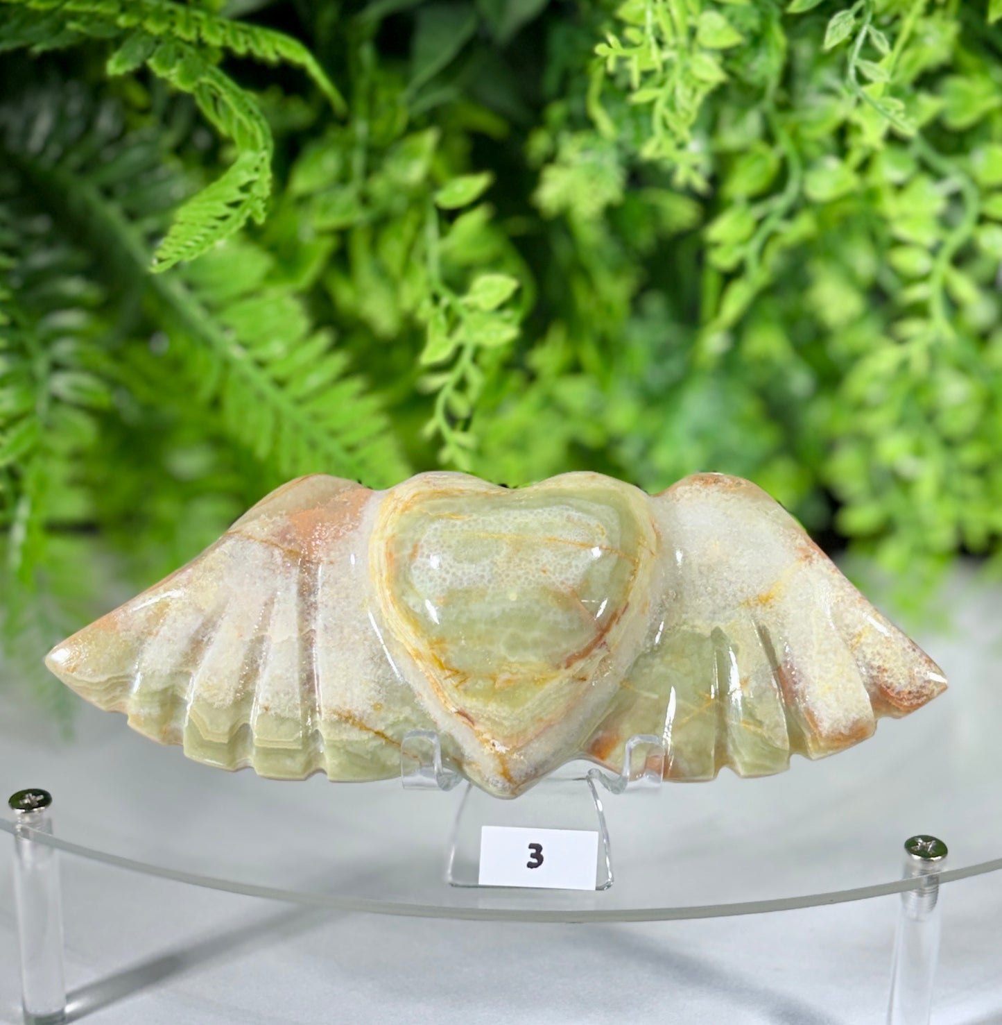 A polished stone winged heart sculpture displayed on a clear acrylic stand, with a small white label showing the number 3. The background features a soft-focus green fern arrangement.