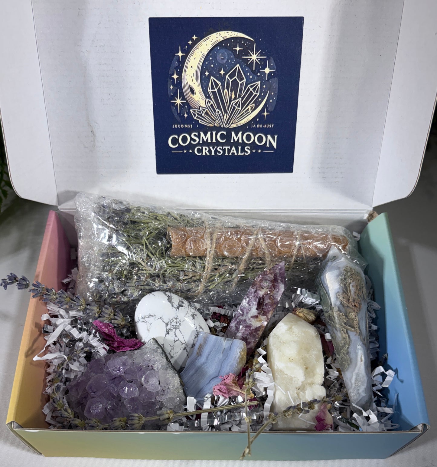 An open gift box from Cosmic Moon Crystals containing a variety of crystals, dried herbs, and a piece of wood on crinkle paper, with the brand logo visible on the inside lid.