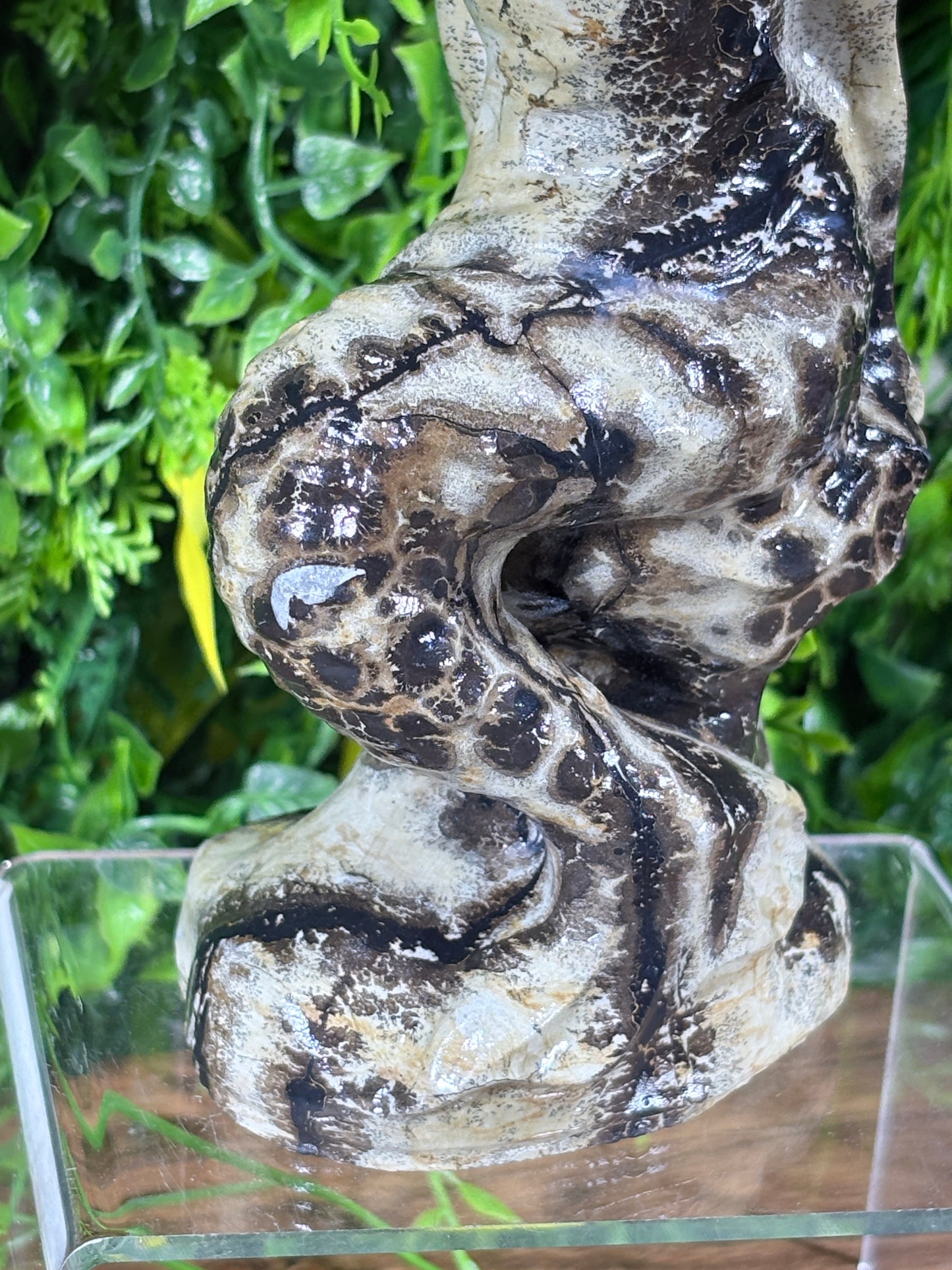 A close-up of a textured, organic-shaped climbing branch or artificial hide element with mottled gray, beige, and black patterning, set against a background of green artificial foliage and a clear enclosure wall.