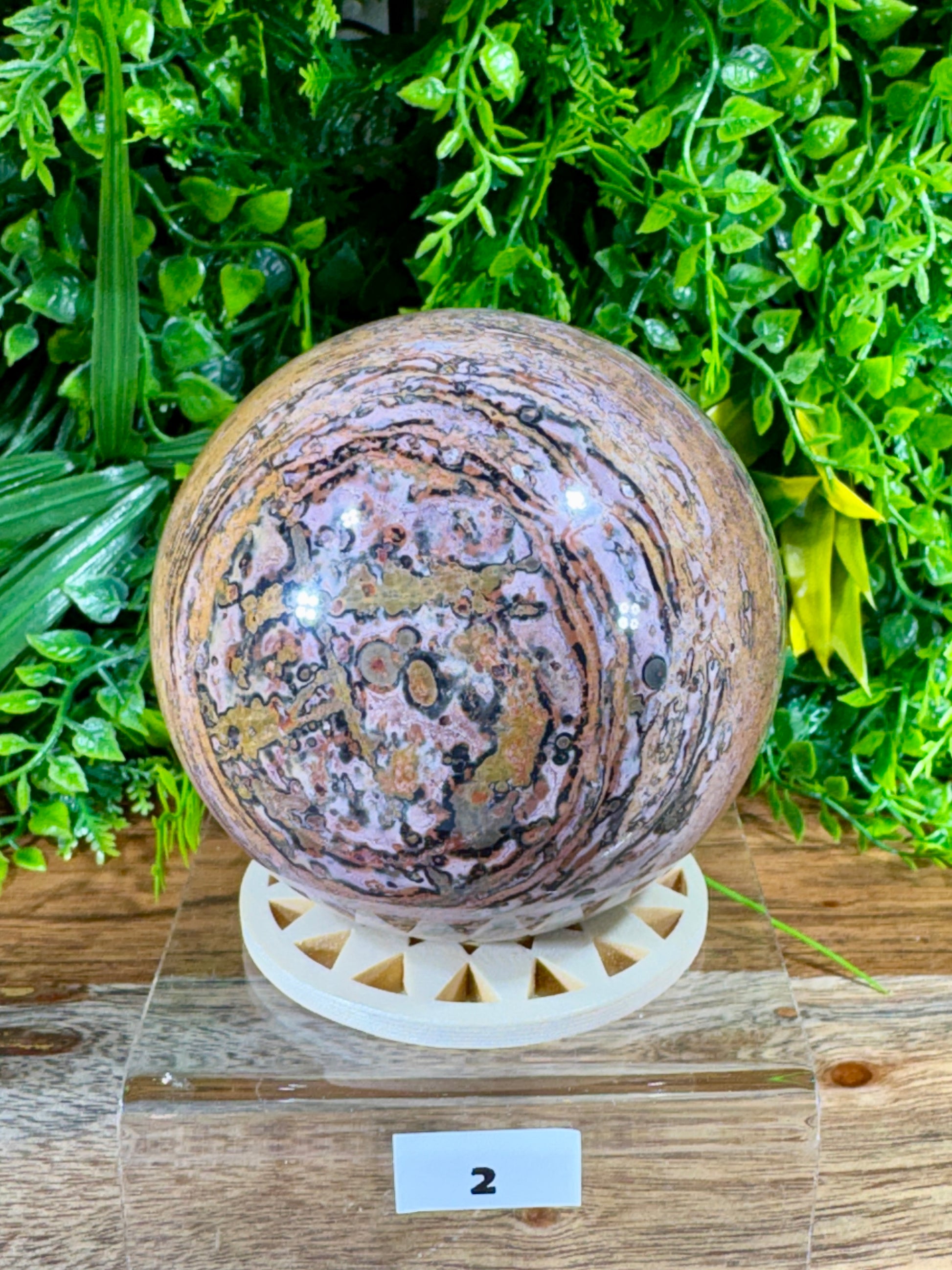 A polished round stone sphere with swirling pink, purple, brown, and gray patterns, displayed on a white circular stand on a wooden surface with green foliage in the background.