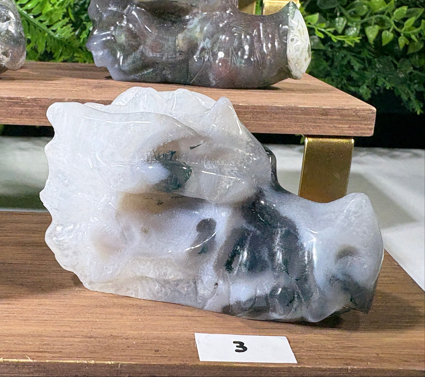 A carved stone sculpture resembling a moss agate dragonm head made from translucent white and gray agate with dark inclusions, displayed on a wooden shelf.
