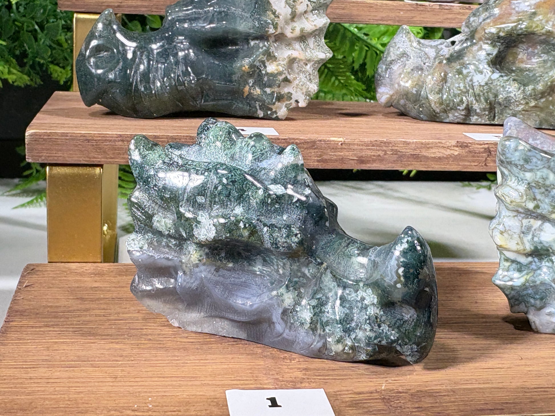 A moss agate polished natural stone carving shaped like a dragon head, displayed on a wooden shelf alongside other raw and finished stone pieces.