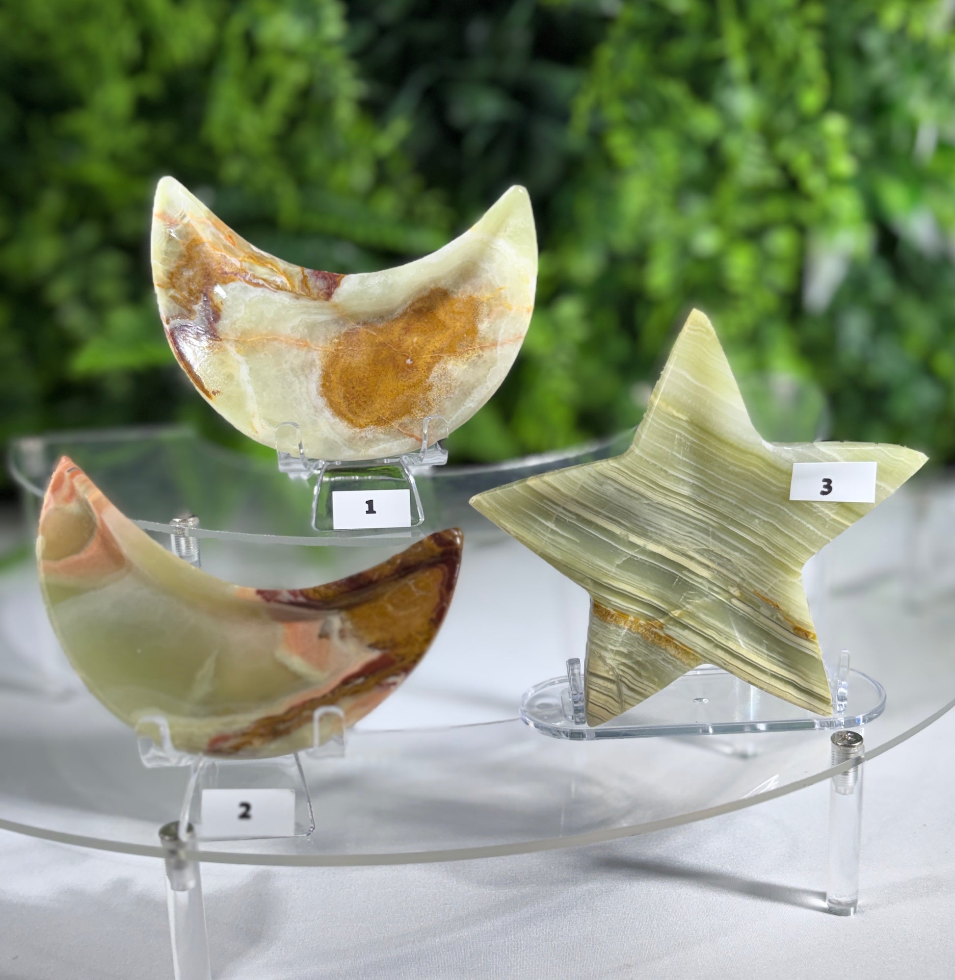 Three polished stone carvings displayed on clear acrylic stands: a crescent moon shape labeled 1, a broken crescent labeled 2, and a star shape labeled 3. The stones show natural marbled patterns in cream, tan, brown, and olive tones, set against a blurred green foliage background.
