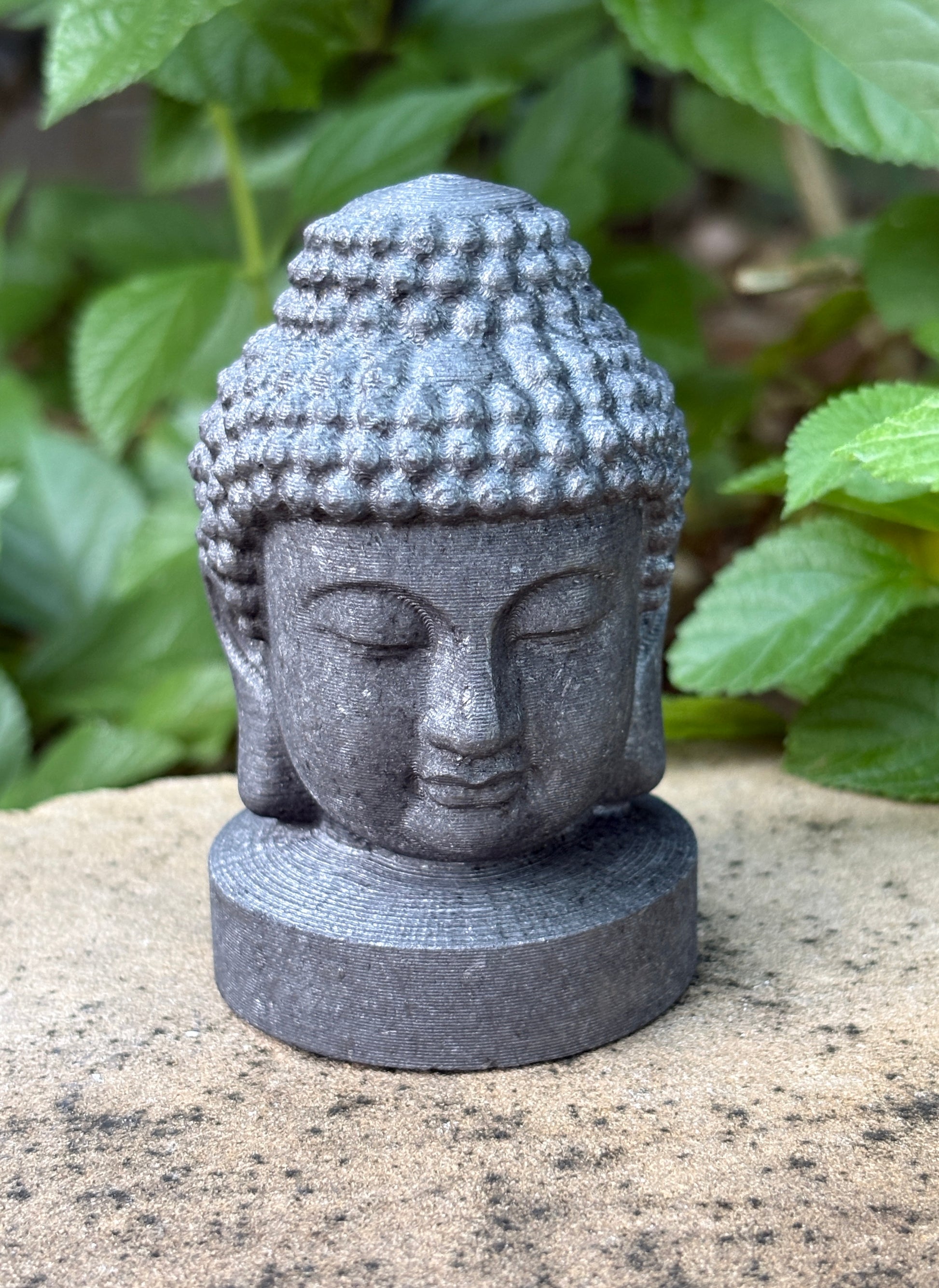 A small gray stone Buddha head statue with closed eyes, standing on a round base outdoors on a sandy surface with green leaves blurred in the background.