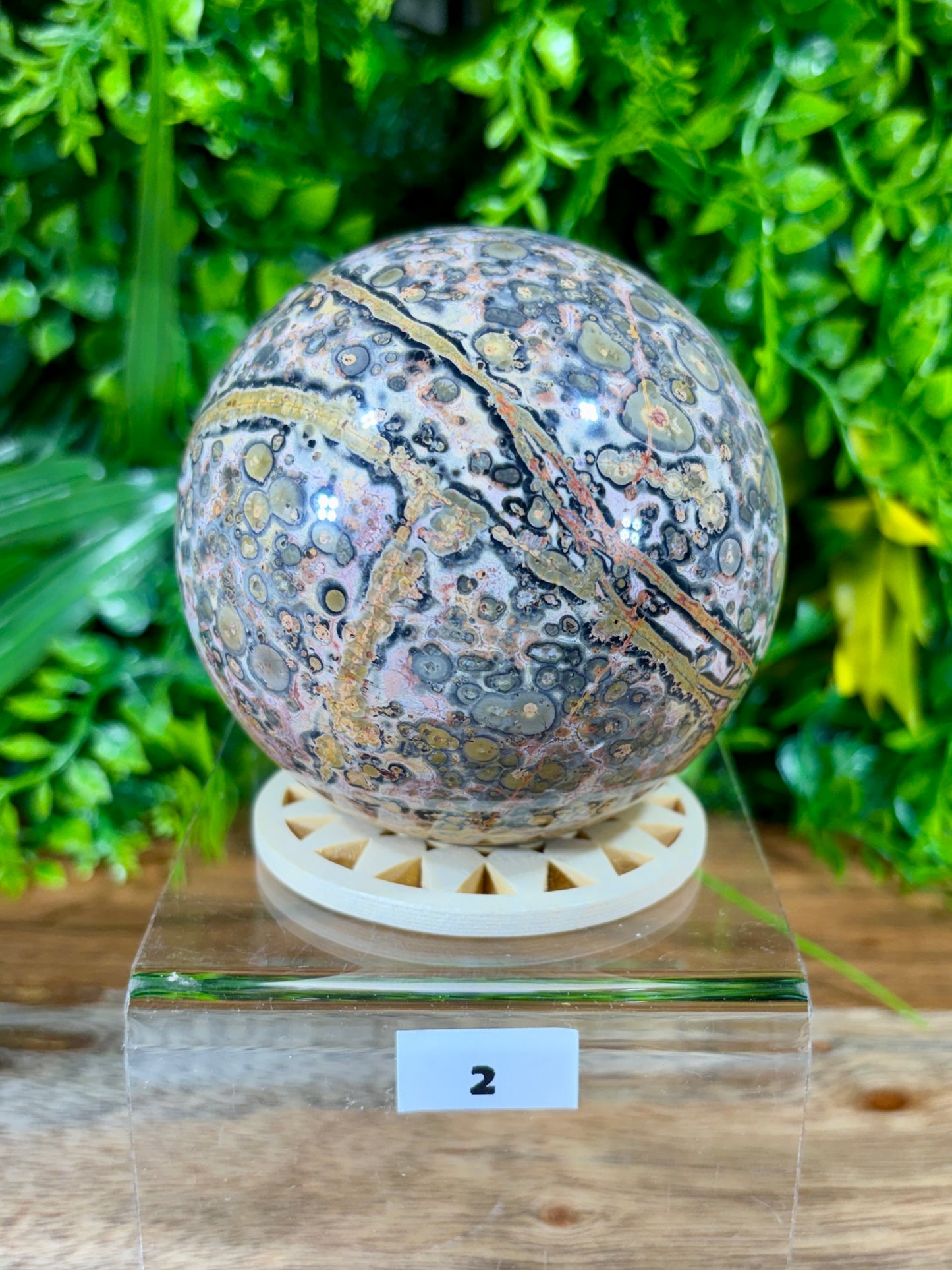A polished round stone sphere with a marbled pattern of gray, gold, pink, and white, displayed on a clear acrylic stand with a small white label showing the number 2. The background consists of artificial green foliage.