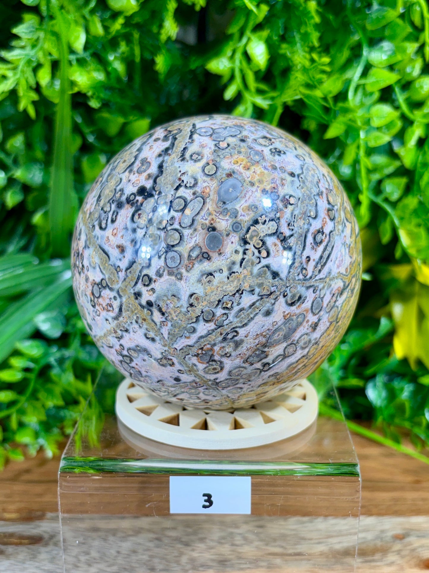 A polished natural stone sphere with intricate swirling patterns of gray, beige, brown, and black, displayed on a small cream-colored circular stand atop a clear acrylic block with a label showing the number 3. The background is a dense, out-of-focus green foliage.