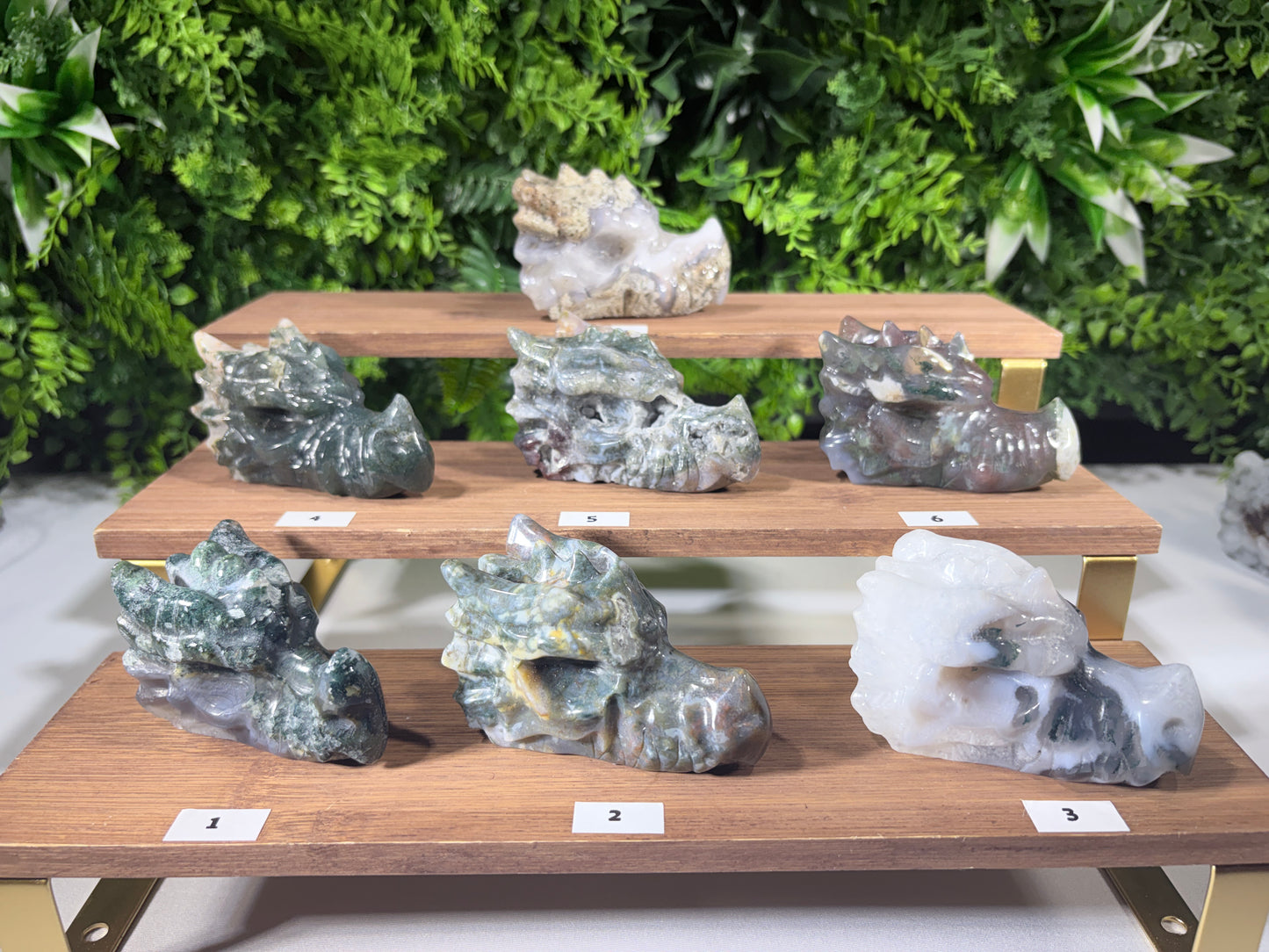 Three wooden display stands holding eight carved stone dragon figurines, numbered 1 through 8 on small white labels, with a green foliage backdrop.