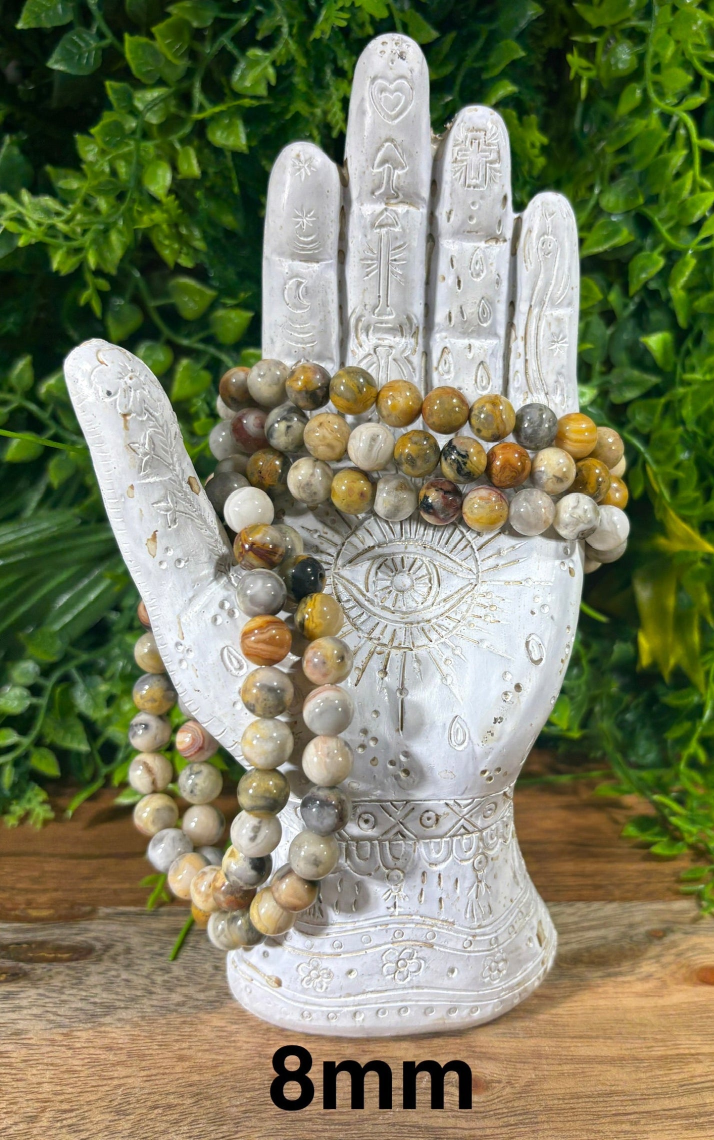 A set of 8mm beaded bracelets with natural stone beads in earthy brown, beige, and gray tones, displayed on a white hand-shaped stand against a green foliage background.