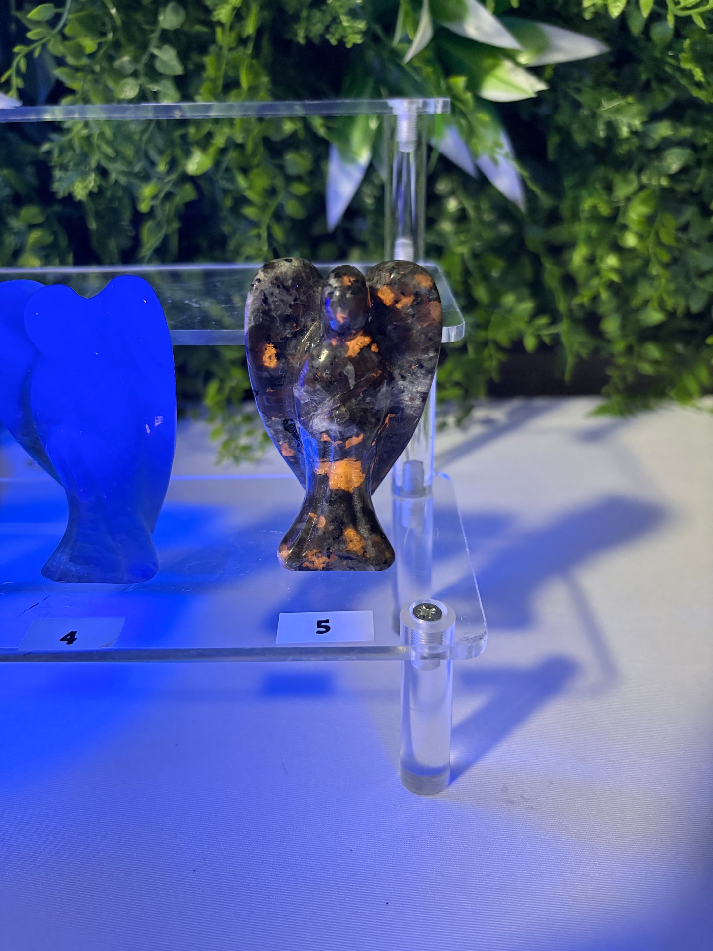A dark brown and black Yooperlight  angel figurine with orange UV inclusions, displayed on a clear acrylic shelf next to a blue glass object, with green foliage in the background.