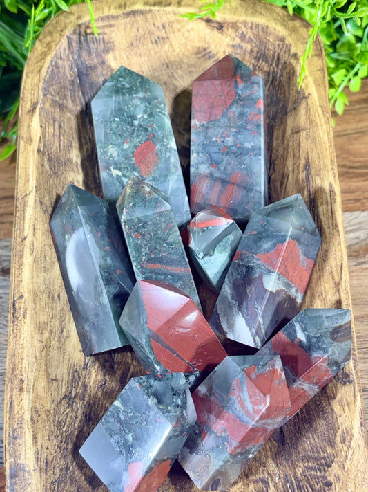 A cluster of polished red jasper crystal points arranged on a rustic wooden tray, with green foliage visible in the background.