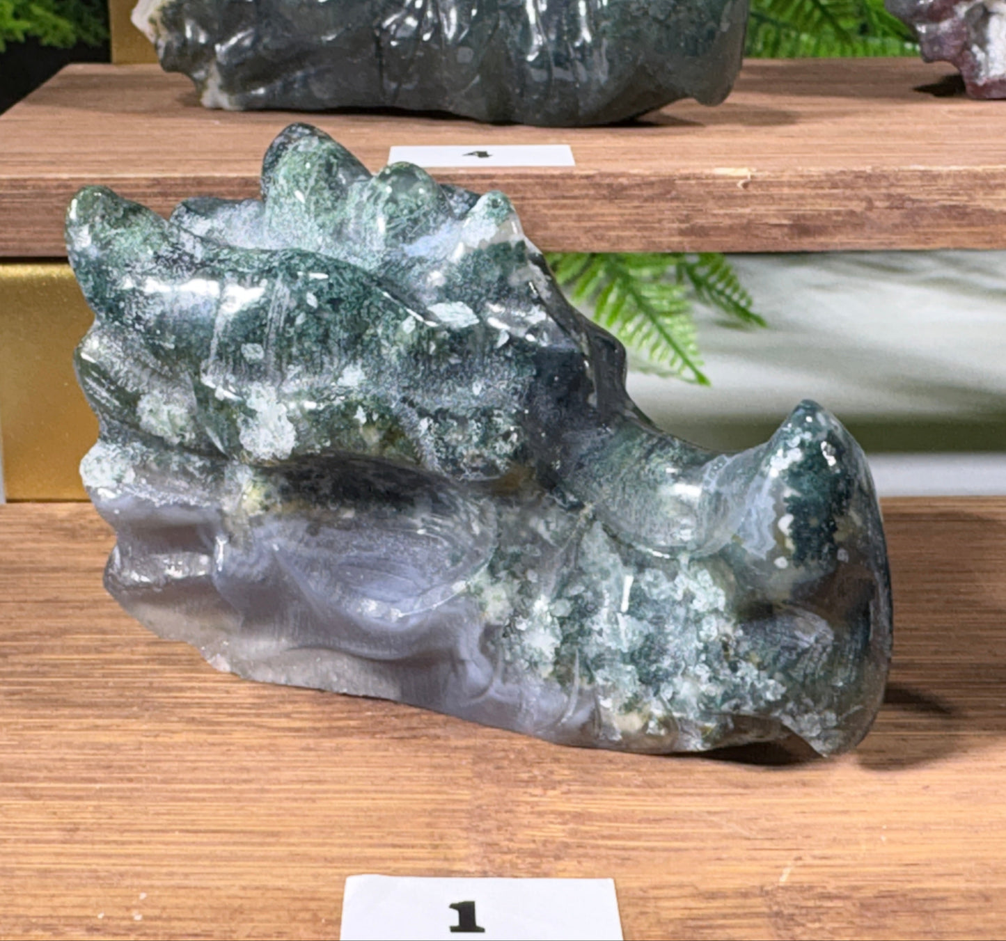 A polished natural moss agate stone dragon carving shaped displayed on a wooden shelf alongside other stones.