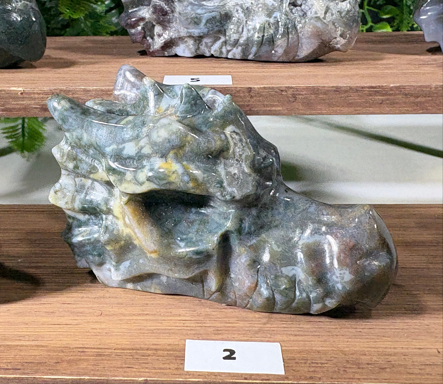 A polished stone sculpture shaped like a dragon head, displayed on a wooden shelf alongside other mineral specimens. The dragon stone has a mottled texture of gray, green, and subtle yellow veins. Moss Agate