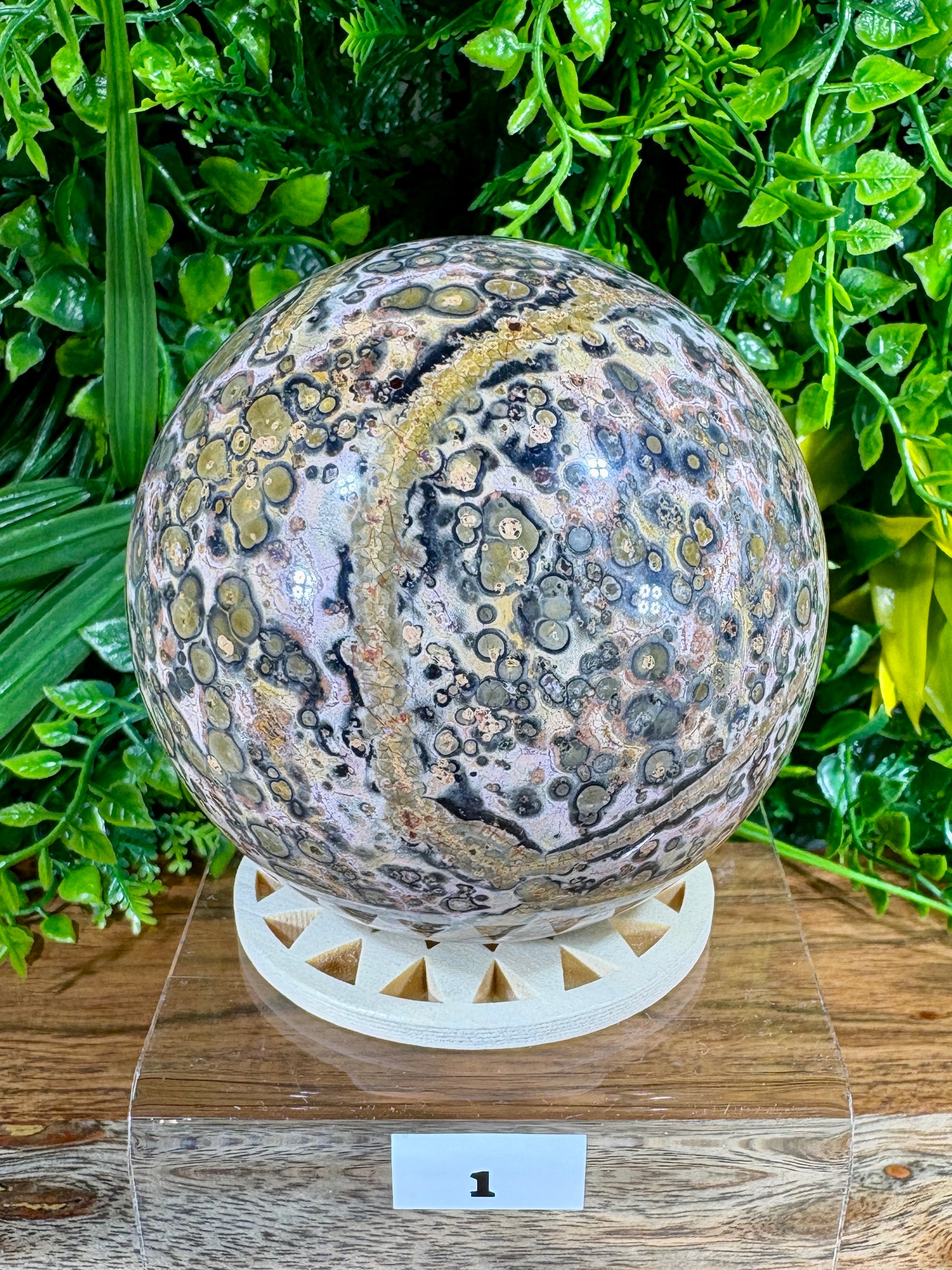 A polished banded stone sphere with pink, gray, black, and gold-toned matrix patterns, displayed on a white geometric stand over a wooden surface with green foliage in the background.