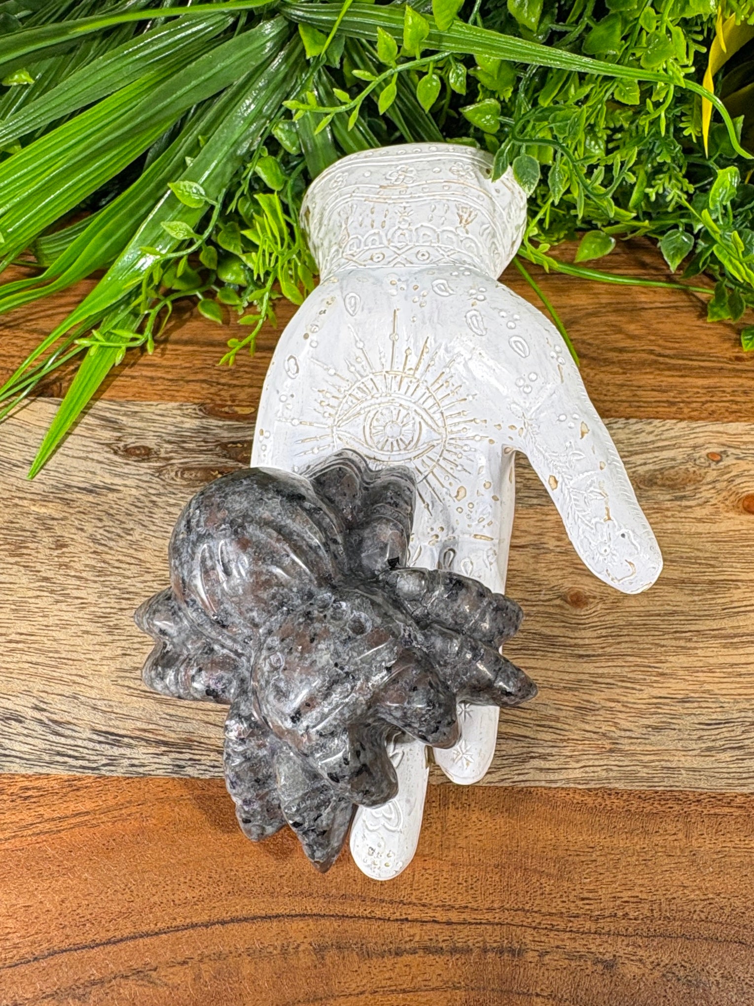 A carved white hamsa hand figurine with gold-toned detailing, holding a dark gray polished crystal cluster, displayed on a wooden surface with green foliage in the background.