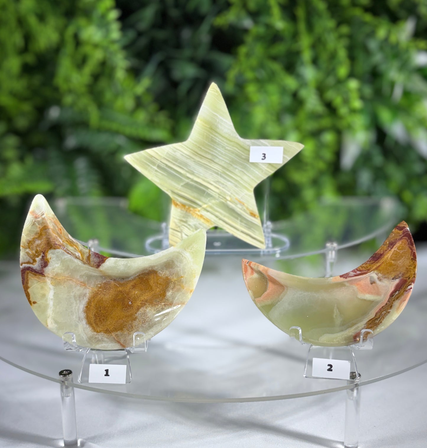 Three polished stone decorative objects displayed on clear acrylic stands: a crescent moon labeled 1, a star labeled 3, and a crescent moon labeled 2, all showing natural marbled patterns in pale green, cream, and brown, set against a blurred green foliage background.
