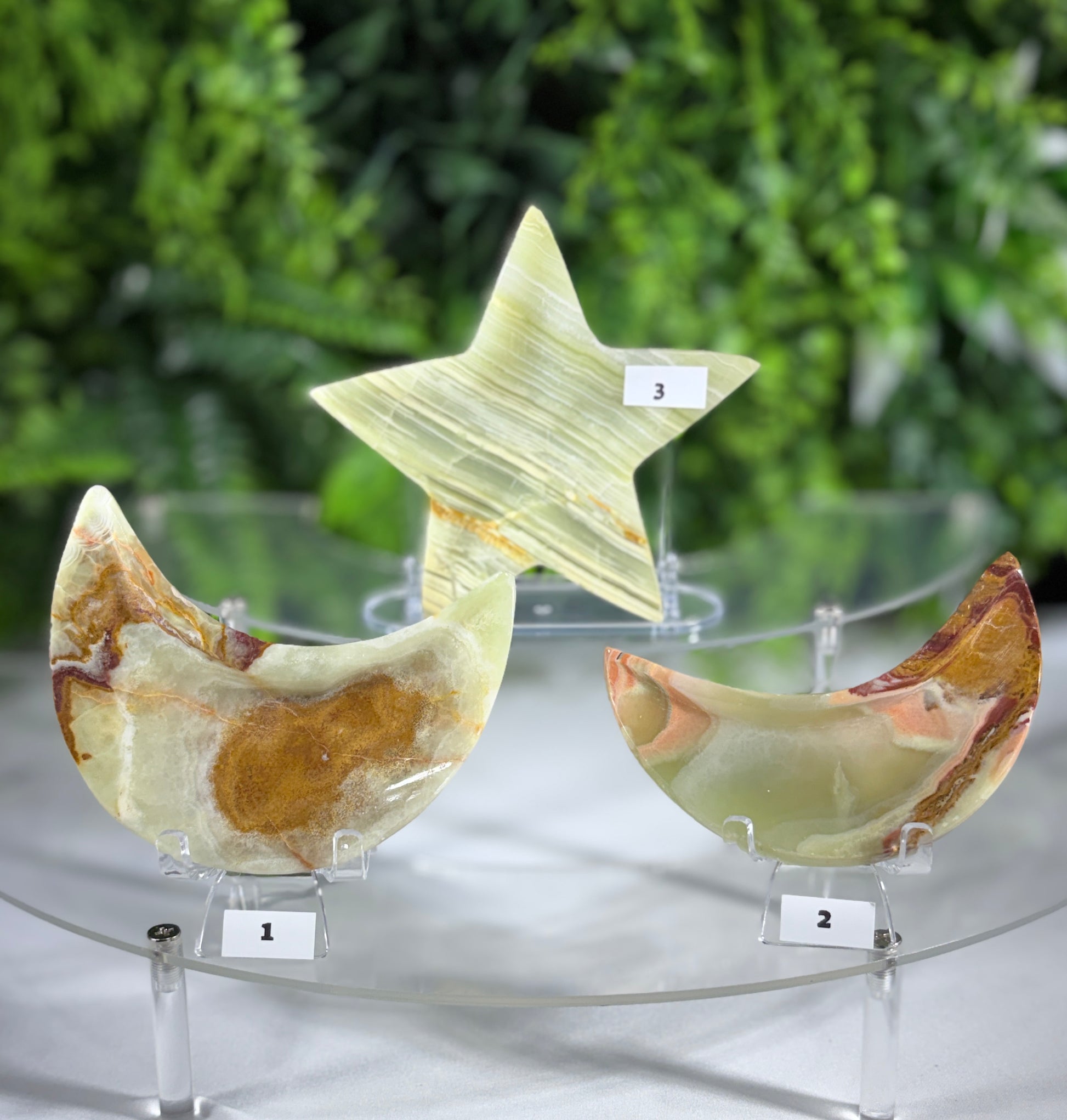 Three polished stone decorative objects displayed on clear acrylic stands: a crescent moon labeled 1, a star labeled 3, and a crescent moon labeled 2, all showing natural marbled patterns in pale green, cream, and brown, set against a blurred green foliage background.