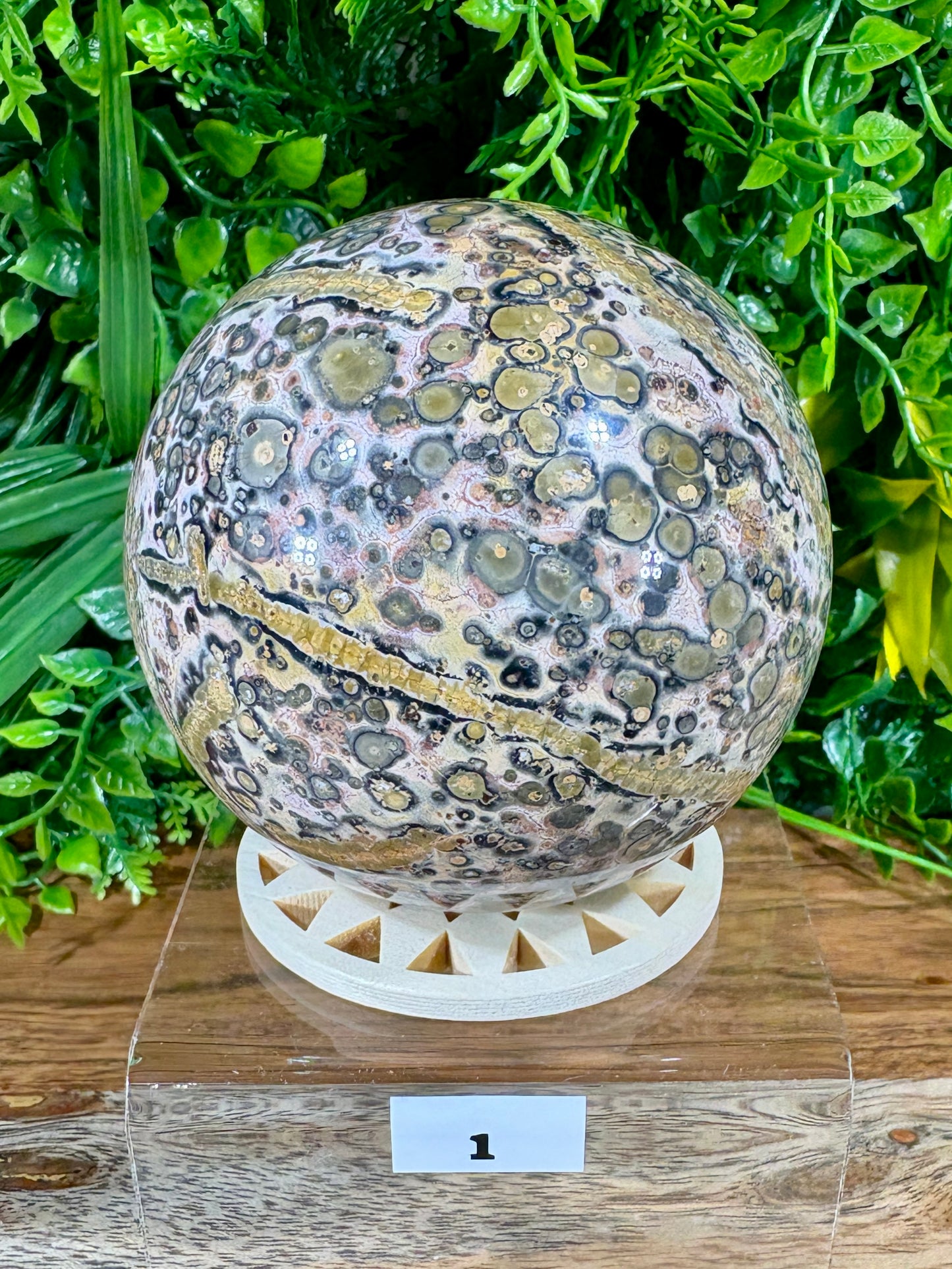 A polished decorative stone sphere with a mottled pattern of beige, gray, black, and gold-like veining, displayed on a white geometric stand over a wooden surface with green foliage in the background.