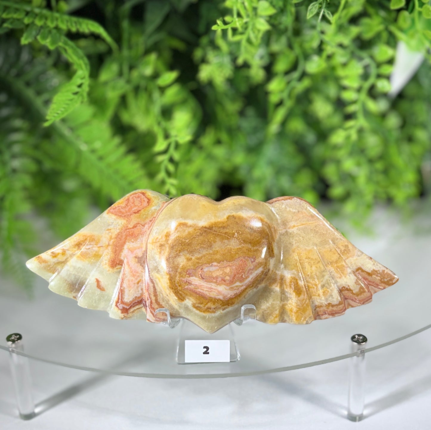 A polished carved stone sculpture of a winged heart, featuring natural marbled patterns in beige, cream, and soft pinkish-brown tones, displayed on a clear acrylic stand with a small label showing the number 2.