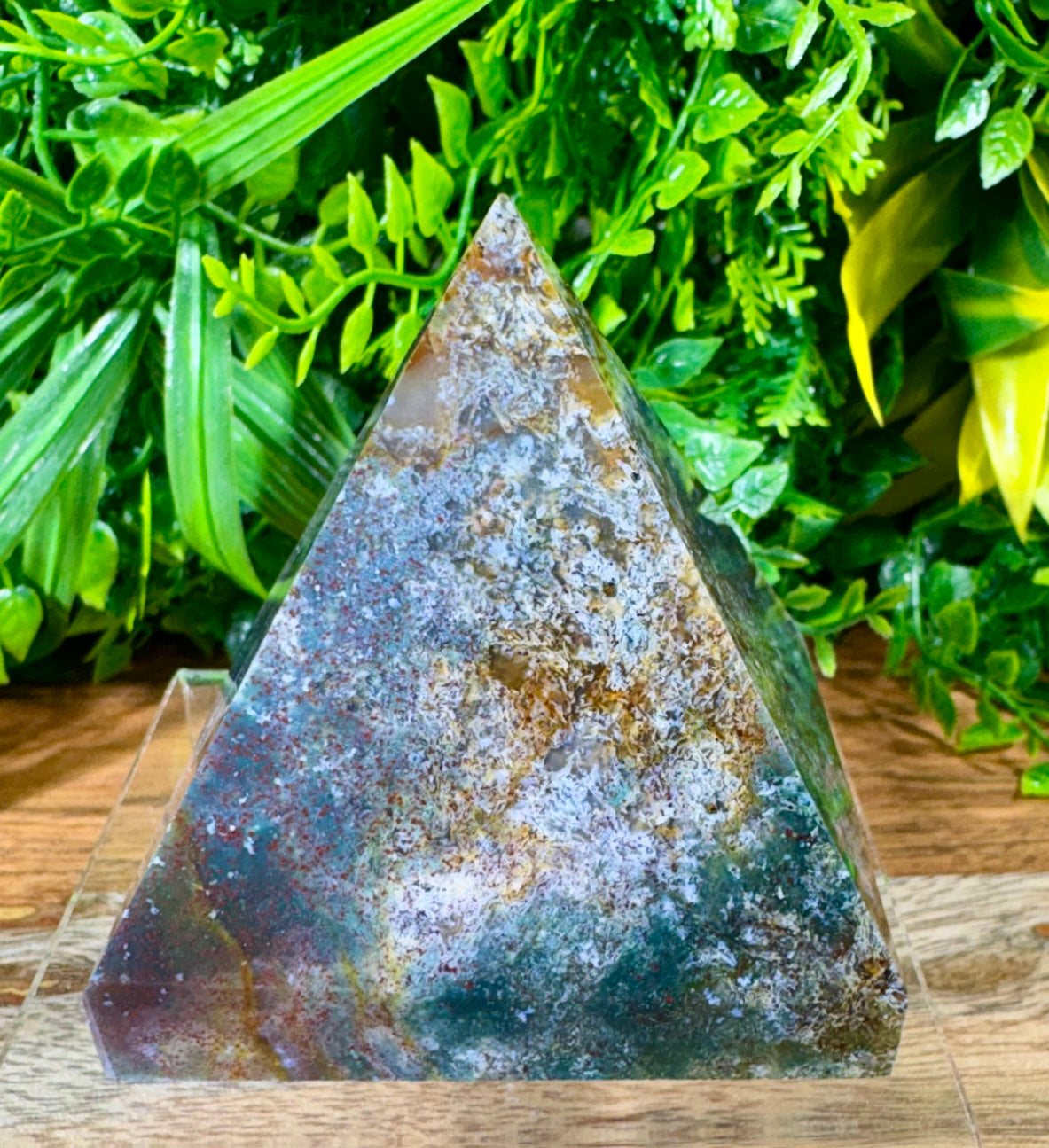 A triangular crystal pyramid with a mottled, metallic appearance in silver, bronze, and dark green, displayed on a clear acrylic stand against a background of artificial green foliage.