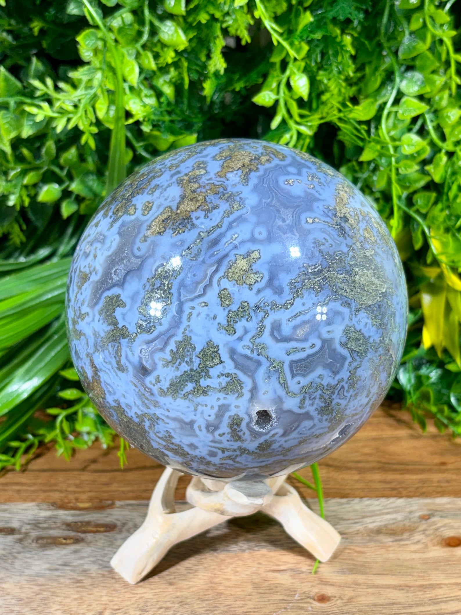 A polished blue and green agate crystal sphere with  veining, displayed on a small white wooden stand against a background of green foliage and a rustic wooden surface.