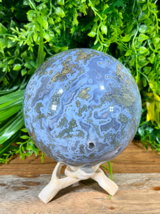 A polished blue and green agate crystal sphere with  veining, displayed on a small white wooden stand against a background of green foliage and a rustic wooden surface.