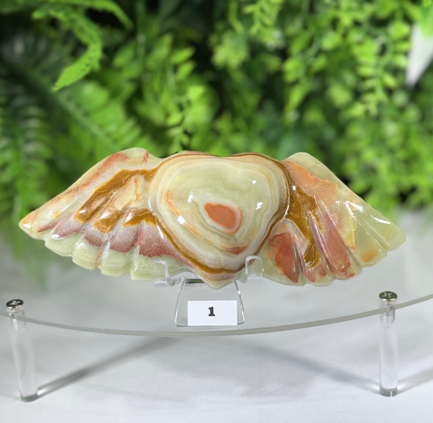 A polished carved stone wing sculpture with swirling cream, tan, and peach bands, displayed on a clear acrylic stand against a blurred green foliage background.