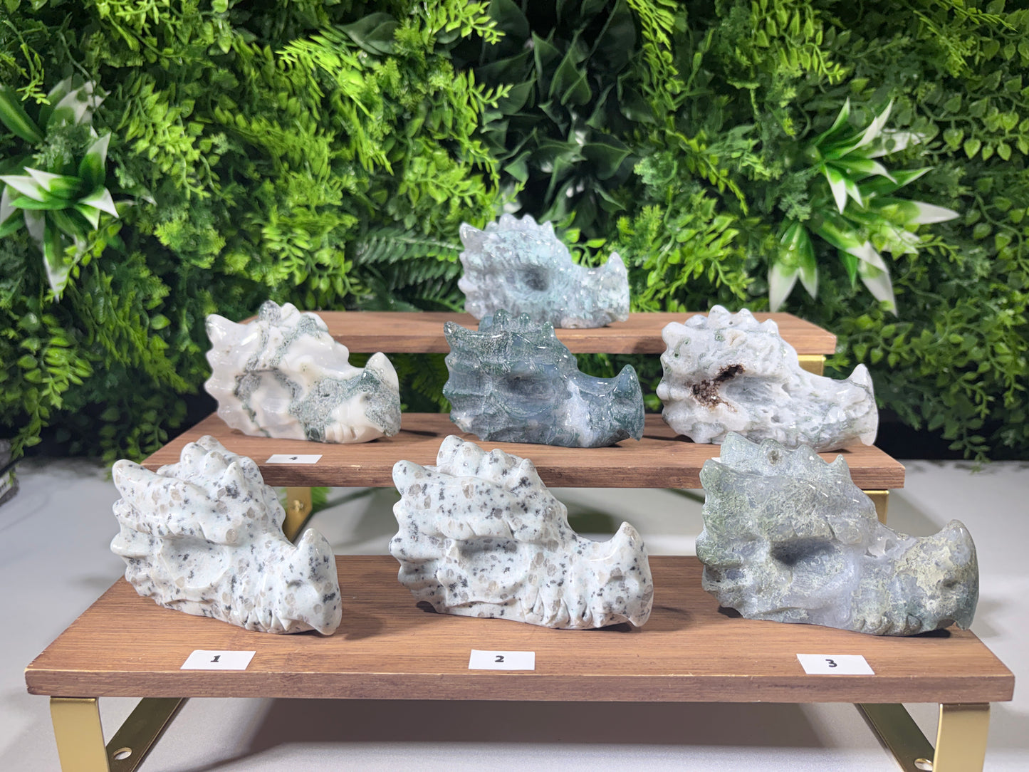 Six natural moss agate and kiwi Jasper Dragon carvings displayed on a wooden tiered stand against a green foliage background. The stones are primarily white and gray with black veining, and some have a blue-gray tint.