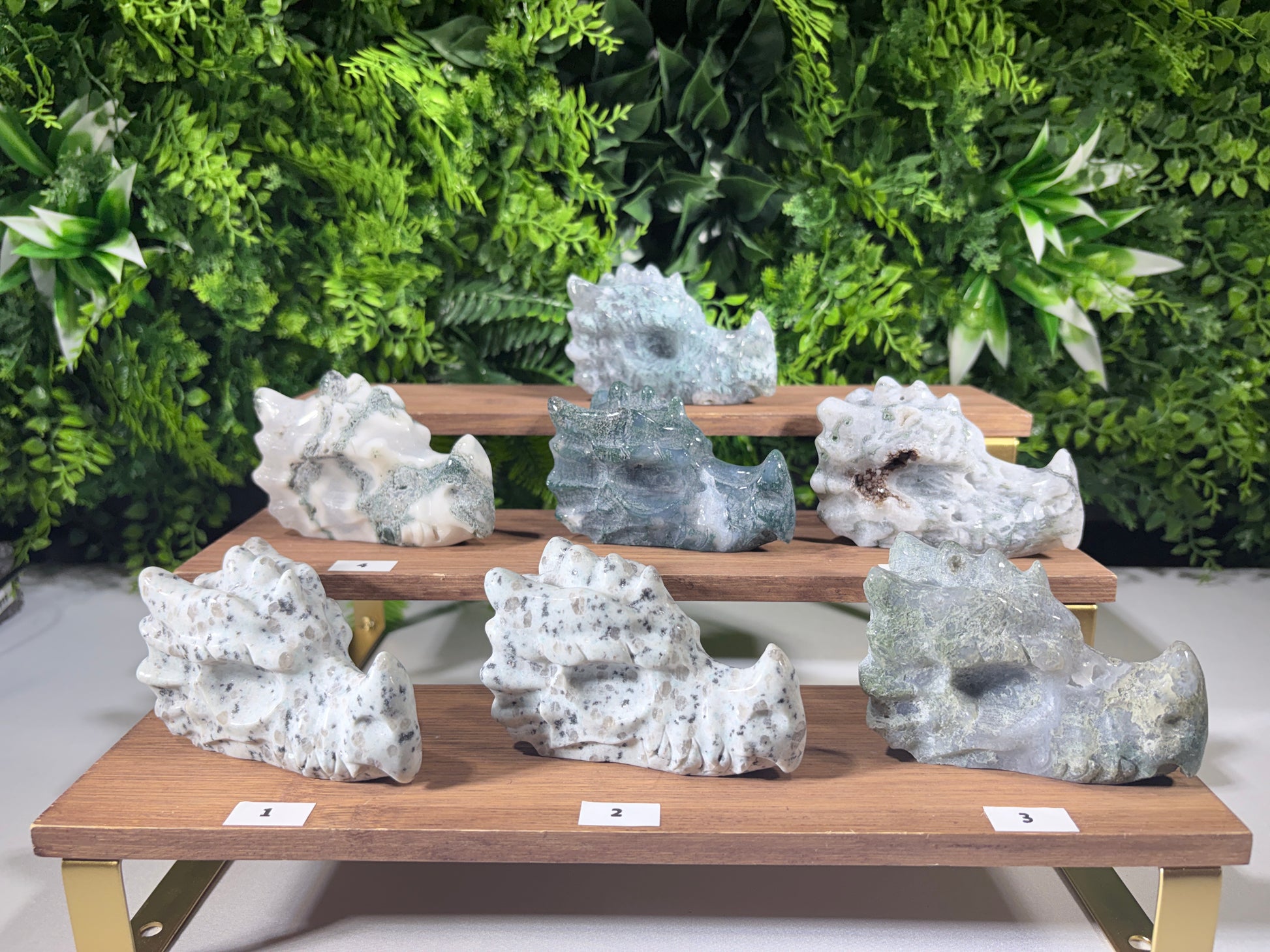 Six natural moss agate and kiwi Jasper Dragon carvings displayed on a wooden tiered stand against a green foliage background. The stones are primarily white and gray with black veining, and some have a blue-gray tint.