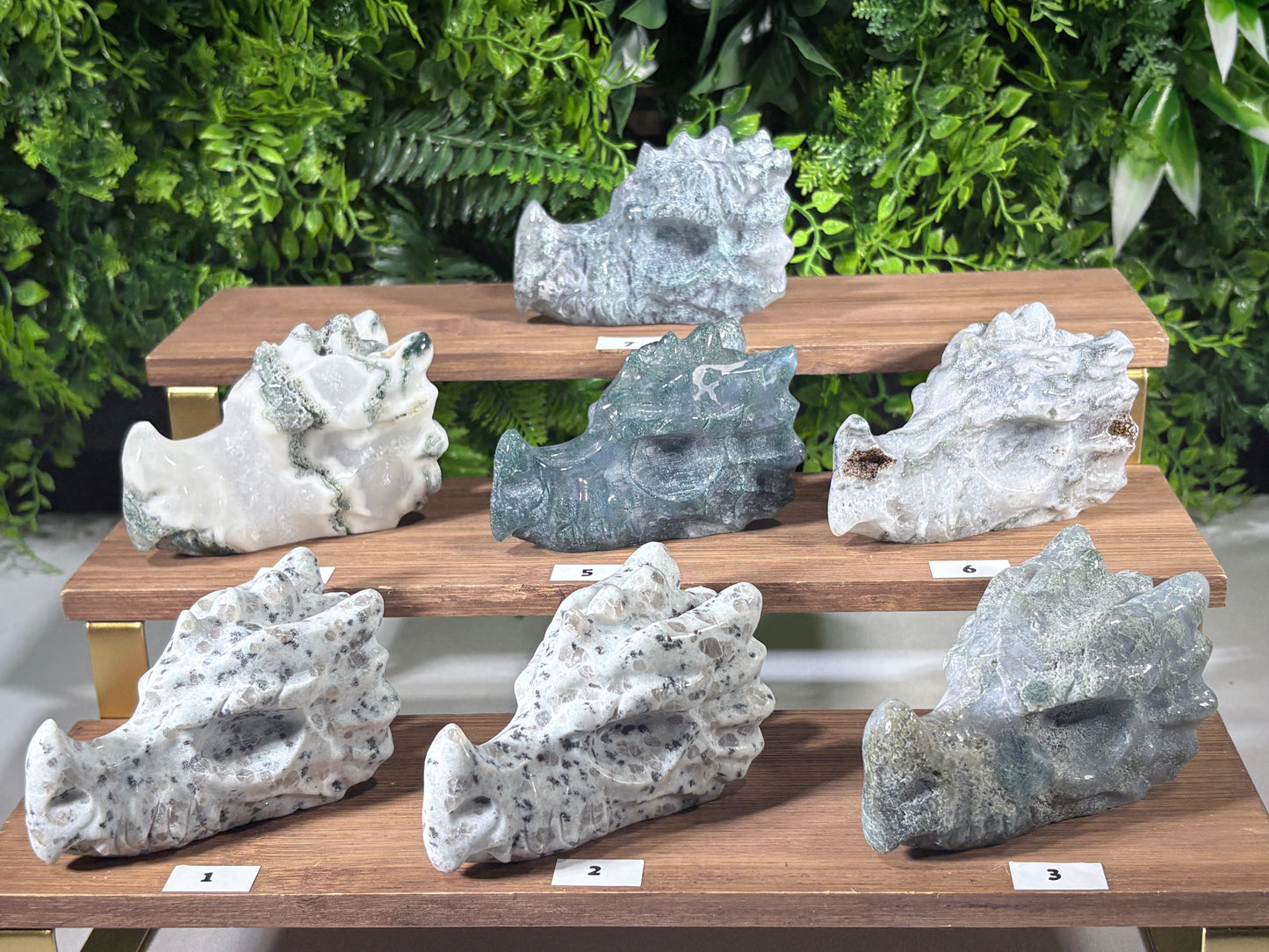 Eight natural stone moss agate and kiwi jasper carved dragon sculptures of various shapes and colors displayed on three wooden tiers against a green foliage background.