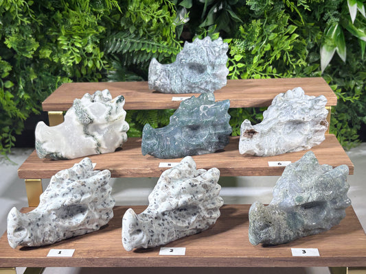 Eight natural stone moss agate and kiwi jasper carved dragon sculptures of various shapes and colors displayed on three wooden tiers against a green foliage background.