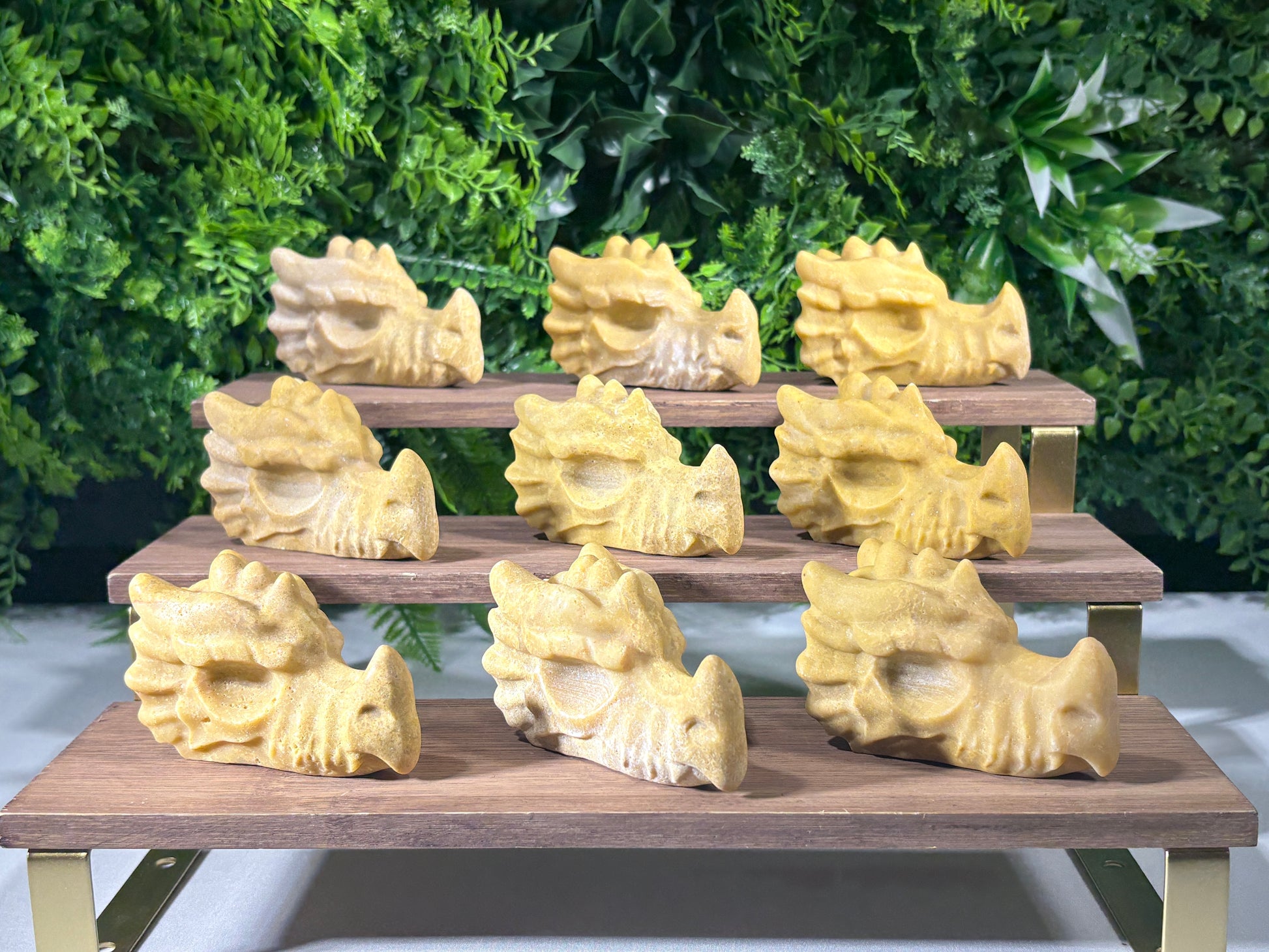 A set of nine pale yellow jade carved dragon heads arranged on a wooden display rack with gold metal brackets, set against a white wall and a green foliage backdrop.