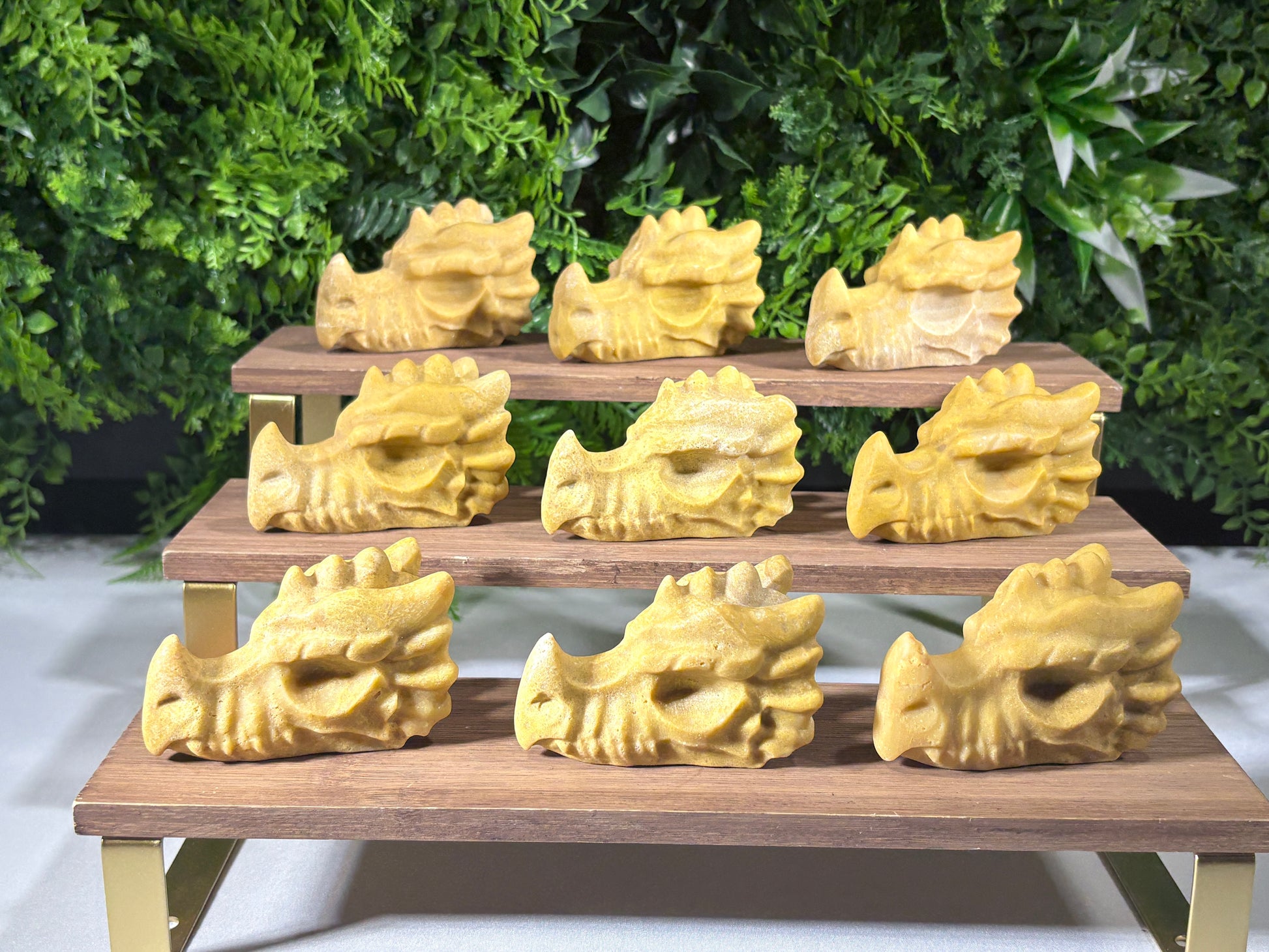 A set of nine pale yellow jade carved dragon heads arranged on a wooden display rack with gold metal brackets, set against a white wall and a green foliage backdrop.