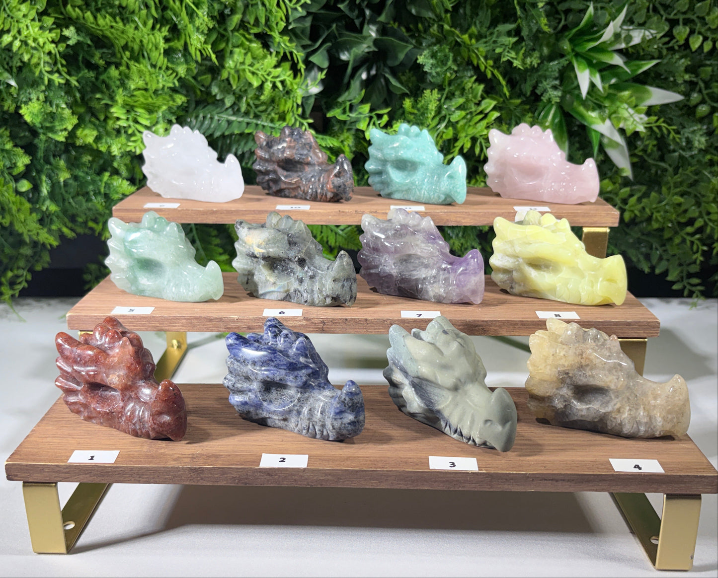 A set of 12 carved crystal dragon figurines in various colors, arranged on a three-tier wooden display stand with small white labels in front of each. The background is a green leafy wall.