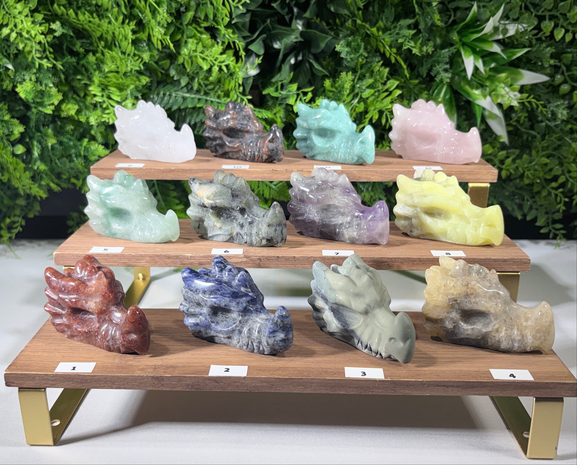 A set of 12 carved crystal dragon figurines in various colors, arranged on a three-tier wooden display stand with small white labels in front of each. The background is a green leafy wall.