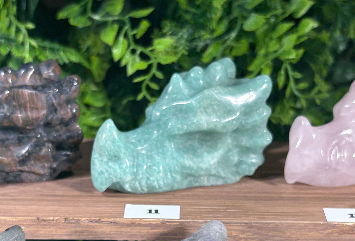 A carved crystal dragon made of purple amazonite sits on a wooden display shelf, surrounded by other colorful crystal formations including aquamarine, pink, gray, and pale yellow stones.
