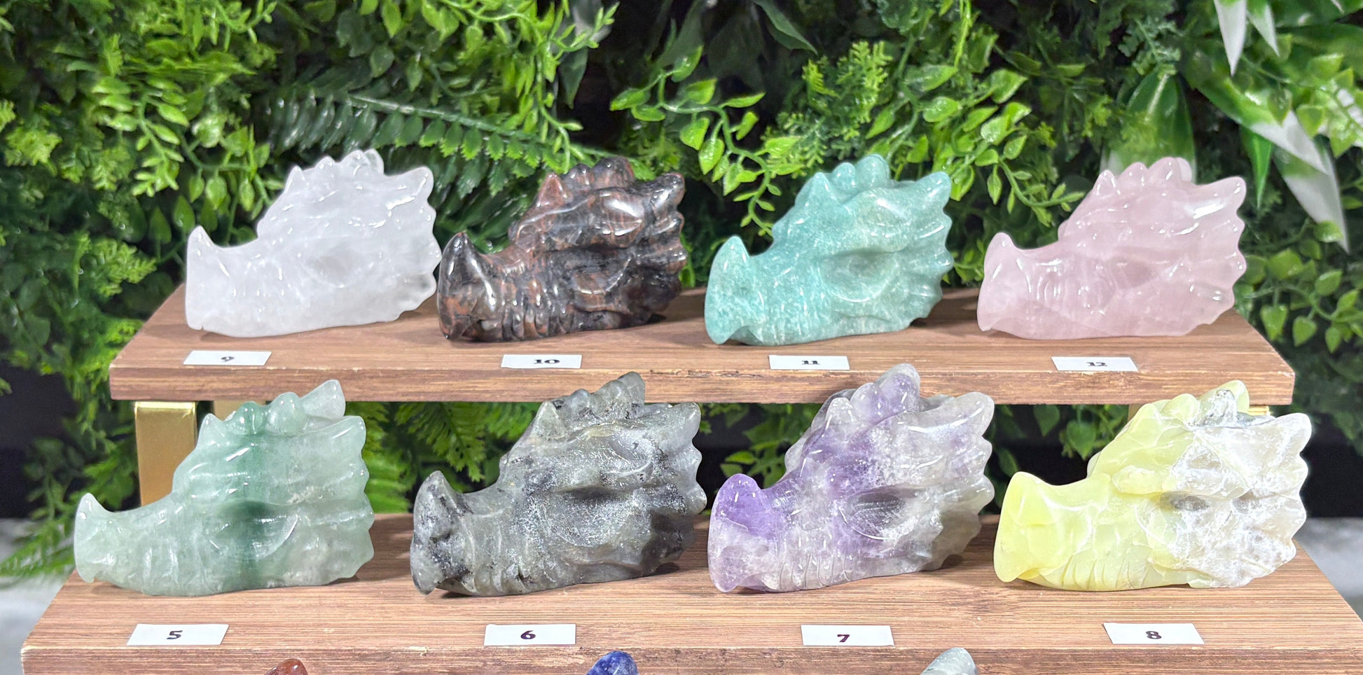 Three wooden display stands holding multiple polished crystal dragon figurines in various colors, set against a green foliage backdrop.