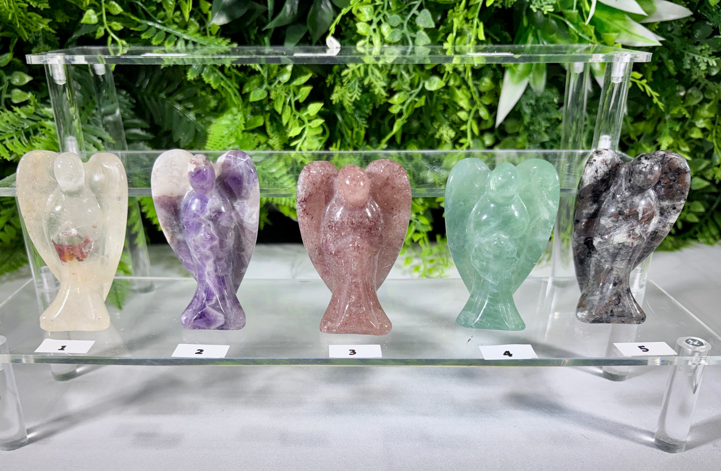 Five carved crystal angel figurines in different colors are lined up on a clear acrylic display shelf against a background of green foliage. The figurines are made of stone with visible grain, and each has a small white label with a number underneath it.