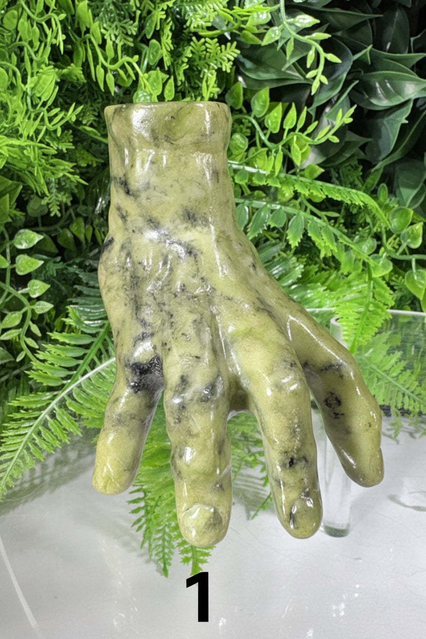 A sculptural hand-shaped vase made of polished olive-green stone with dark marbled inclusions, displayed against a backdrop of artificial green foliage.