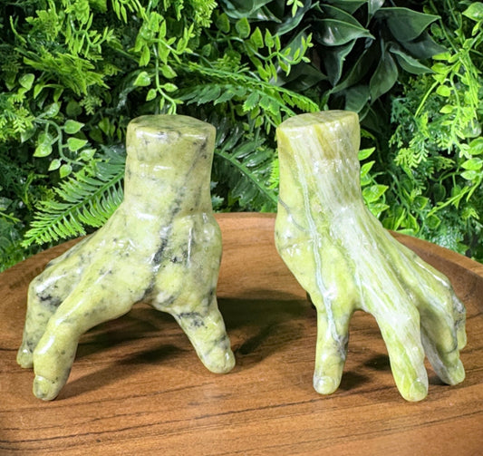 Two green stone claw sculptures displayed on a wooden surface in front of a green foliage backdrop.