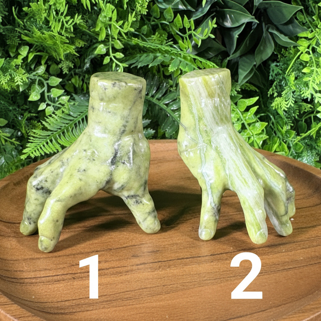 Two green stone claw sculptures displayed on a wooden surface in front of a green foliage backdrop.