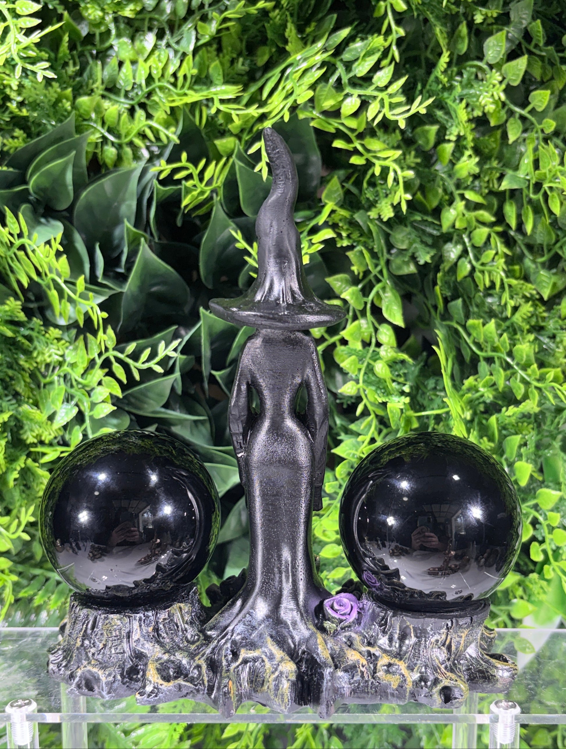 A black witch figurine with a tall pointed hat, flanked by two glossy black spheres on a textured base, displayed against a green leaf backdrop.