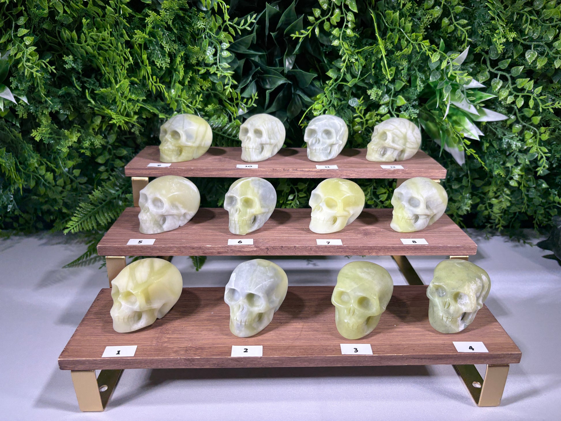 Three wooden display shelves arranged in a stepped pyramid, each holding multiple carved skull sculptures in pale yellow, green, and gray stone. Small white labels with numbers are placed under the shelves, and a green leafy backdrop fills the background.