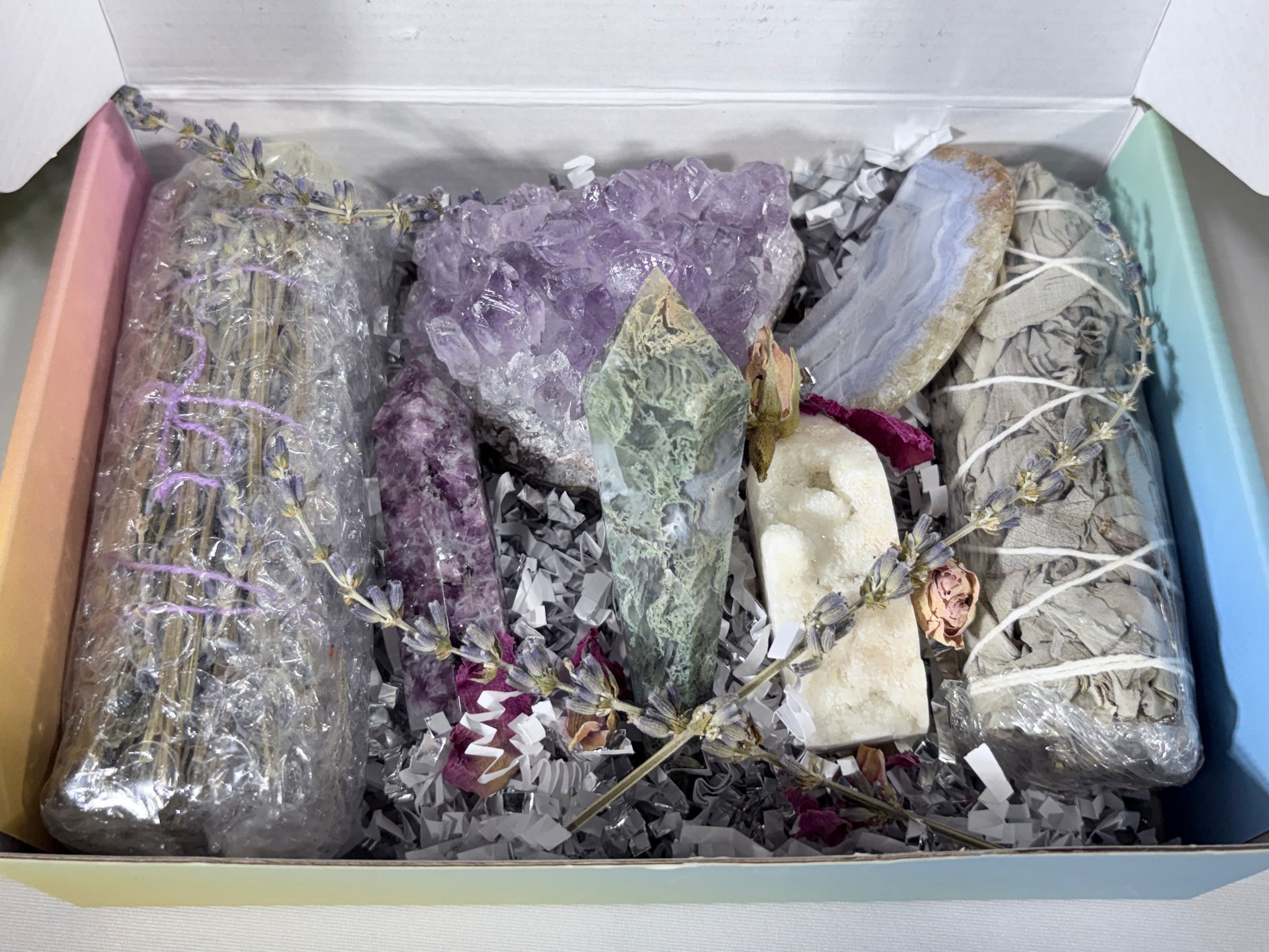 A gift box filled with natural crystals, dried lavender bundles wrapped in clear plastic, a white agate slice, and white sage smudge sticks on shredded paper inside a pastel-colored box.
