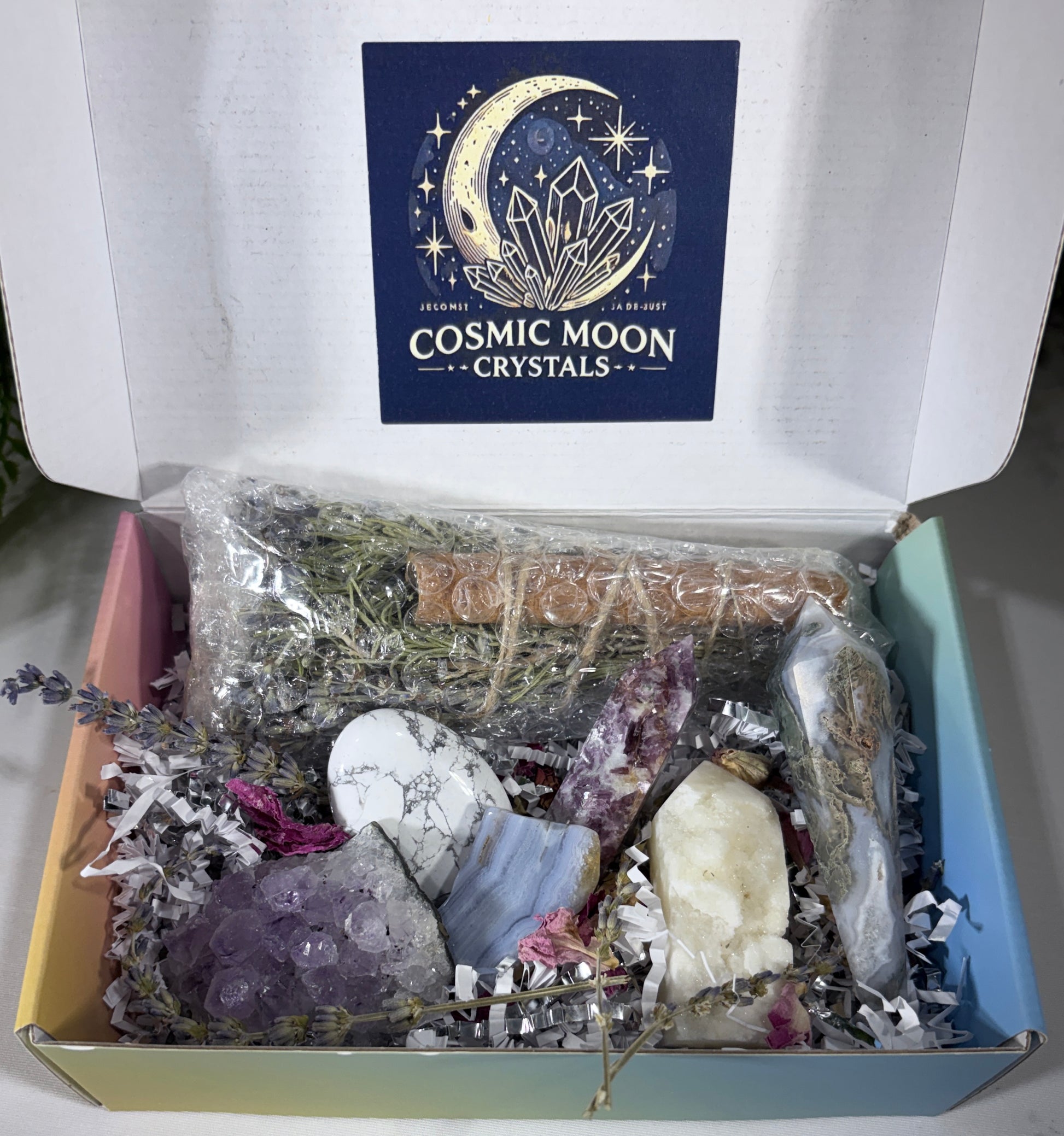 An open gift box from Cosmic Moon Crystals containing a variety of crystals, dried herbs, and a piece of wood on crinkle paper, with the brand logo visible on the inside lid.