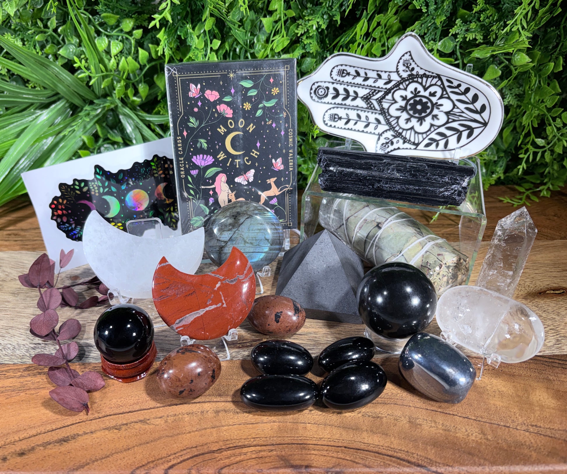 A collection of crystals, stones, and decorative items arranged on a wooden surface with green foliage in the background. The set includes black obsidian stones, a red-orange stone, clear quartz points, a white hamsa hand print, a black-and-white patterned card, a colorful card, and several smaller decorative pieces.