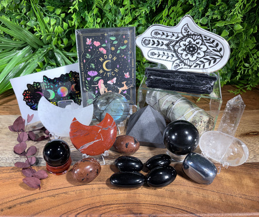 A collection of crystals, stones, and decorative items arranged on a wooden surface with green foliage in the background. The set includes black obsidian stones, a red-orange stone, clear quartz points, a white hamsa hand print, a black-and-white patterned card, a colorful card, and several smaller decorative pieces.