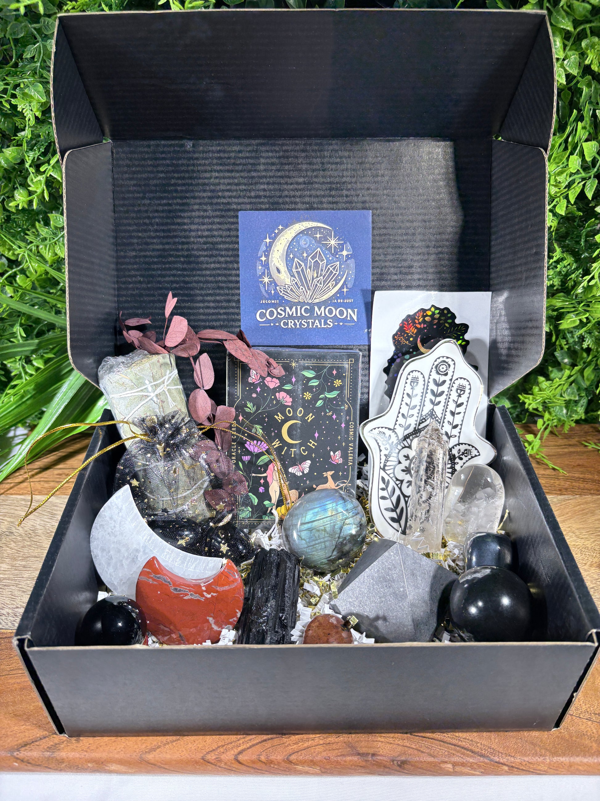 A black gift box filled with crystals, stones, a tarot card deck, a Hamsa hand charm, and a blue card reading 'Cosmic Moon Crystals,' set against a background of green foliage.