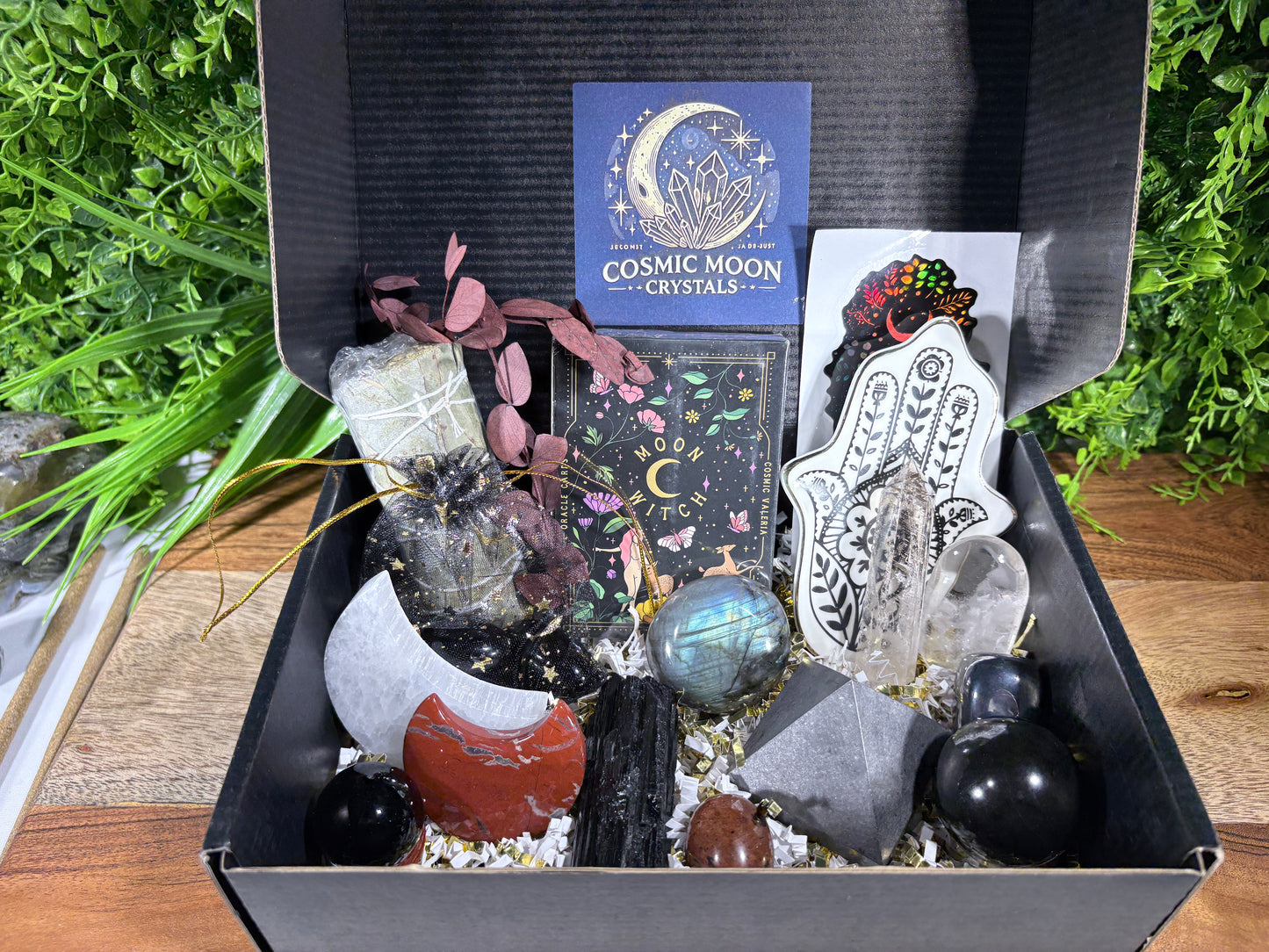 A curated crystal and moon-themed gift box set displayed inside an open black box. Visible items include a blue card reading 'COSMIC MOON CRYSTALS,' a white Hamsa wall decor piece, a clear crystal cluster, black and red polished stones, a gray stone, a small green crystal, and decorative dried leaves and a black cord.
