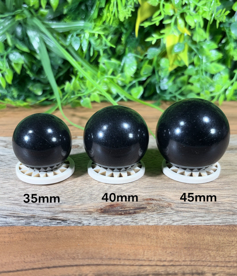 Three glossy black spherical stone ornaments displayed on small white geometric stands, arranged in a row on a wooden surface with a blurred green foliage background.