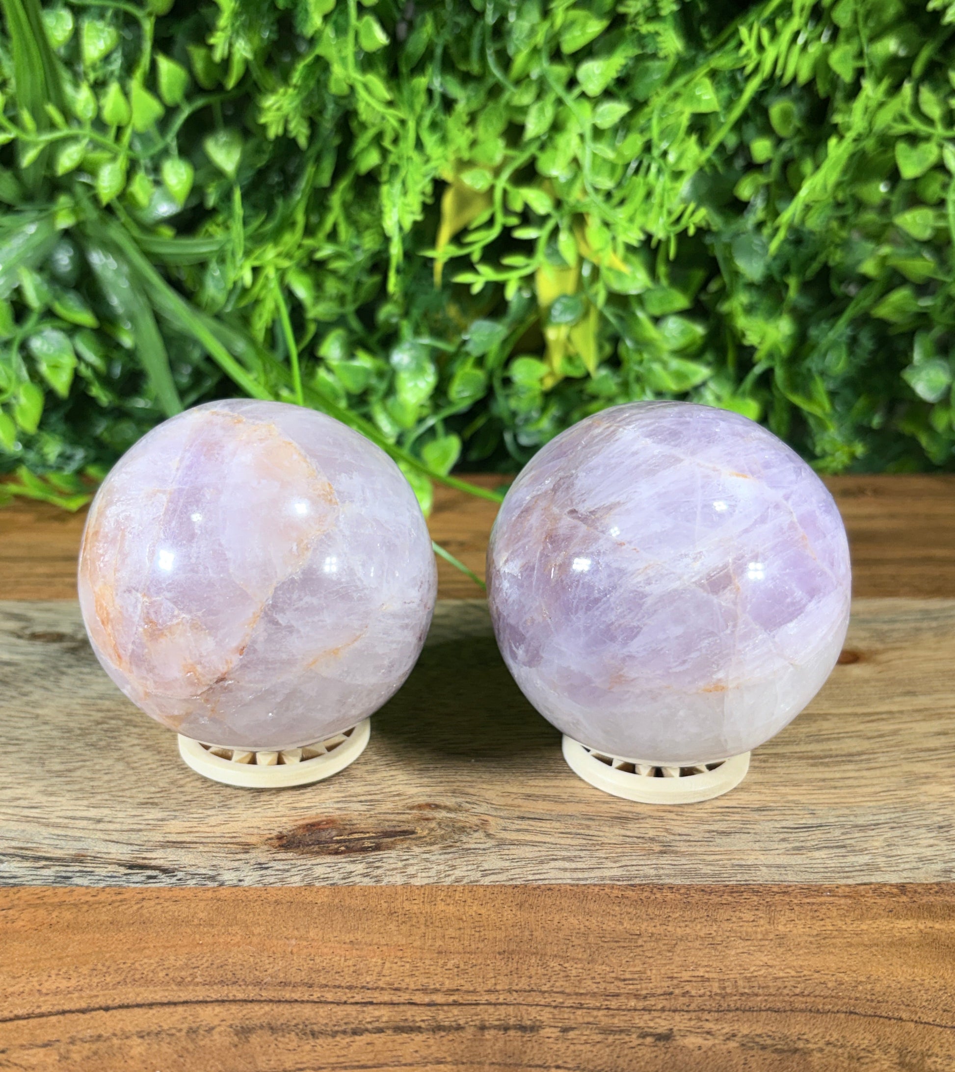 Two round lavender quartz crystal spheres with cream-colored metal wall mounts, displayed vertically against a rustic wooden panel next to a green foliage background.